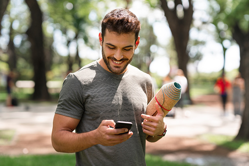 A muscular young man with an exercise mat under his arm is sending text messages with his smart phone.