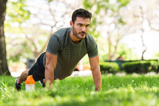 A handsome young man doing push-ups in the park. He has a protein drink right next to him.