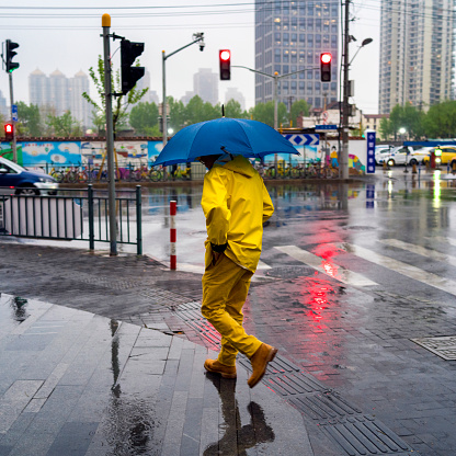 Shanghai, China - April 13th, 2018 : Man wearing a yellow rain suit and holding a blue umbrella crossing the street in rain.