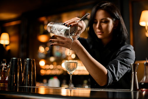 Professional bartender girl pouring a trasparent alcoholic drink from the measuring cup to the glass through the strainer filter on the bar counter