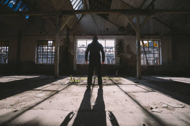 Adult man standing inside some large, dark, spooky,abandoned building illuminated with sunlight through window Photo is taken with full frame dslr camera indoors. serial killings photos stock pictures, royalty-free photos & images