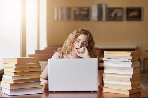 Portrait of sleepy hard working sitting at library, closing eyes, yawning, using her laptop during studying, having bunches of books around on desk, spending time at comfortable workingspace.