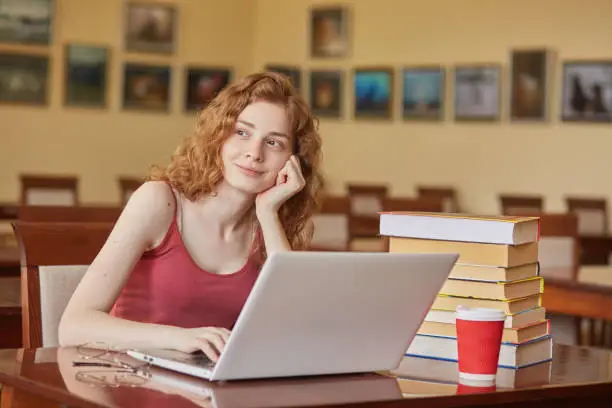 Indoor shot of dreamy adorable young lady looking aside, sitting at table in reading hall, having peaceful facial expression, using laptop while studying, having eyewear and papercup of drink on desk.
