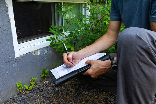 A close up view of a building inspector checking exterior walls and windows of a domestic dwelling, using a pen and paper to fill regulation paperwork.