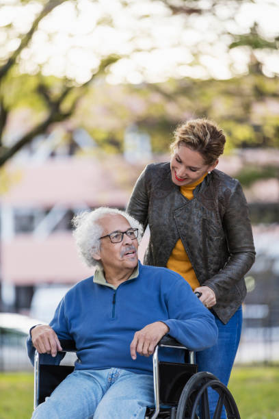 Hispanic woman pushing senior father in wheelchair A senior Hispanic man in his 80s sitting in a wheelchair, talking to his adult daughter, who is in her 30s, pushing the wheelchair from behind. They are walking in a city park in fall, building out of focus in the background. father daughter 80s stock pictures, royalty-free photos & images