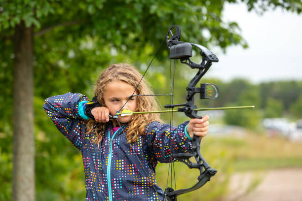 Close-up of a young girl (nine years old) practicing shooting her bow and arrow at an outdoor archery range on a fall day. The girl has her arrow drawn and is aiming at a target - not visible in this image.