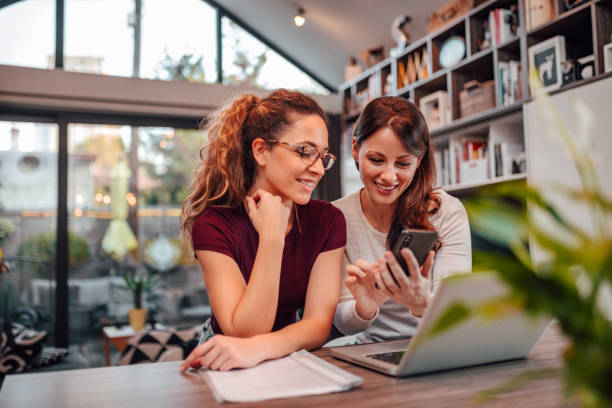 Two smiling woman looking at smart phone while sitting a desk with laptop, in modern house, portrait. Two smiling woman looking at smart phone while sitting a desk with laptop, in modern house, portrait. female friendship stock pictures, royalty-free photos & images
