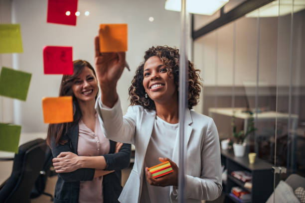 Creative businesswoman writing on sticky notes on a glass wall, female colleague looking. Creative businesswoman writing on sticky notes on a glass wall, female colleague looking. office fun business adhesive note stock pictures, royalty-free photos & images