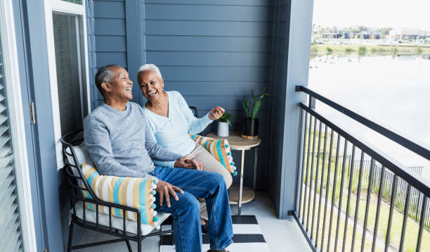 Senior couple relaxing on porch, holding hand, laughing A senior African-American couple sitting side by side on their porch or balcony, relaxing, and holding hands, conversing and laughing together. senior lifestyle stock pictures, royalty-free photos & images
