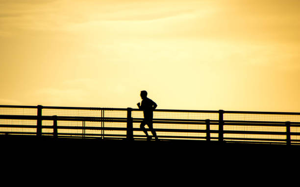 December 24, 2019 : Lincolnshire, England, UK.  An adult male running across a motorway bridge while he tries to improve his health / fitness.  The sunsetting behind him causing the silhouette.