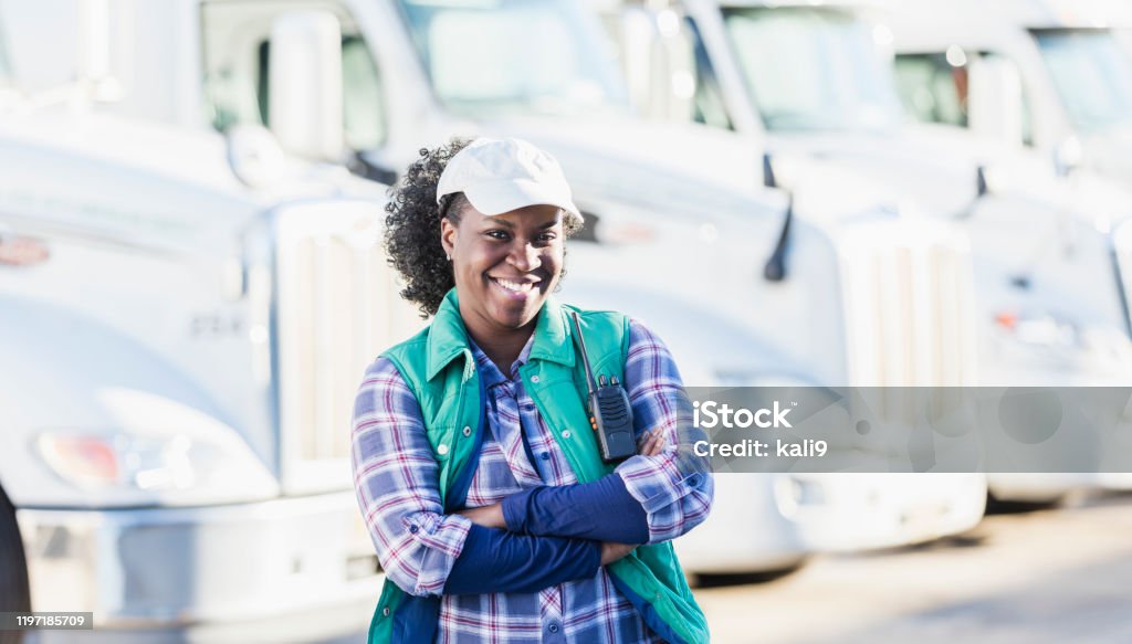 African-American woman standing in front of semi-trucks A mid adult African-American woman in her 30s, a female truck driver standing in front of a fleet of semi-trucks or tractor-trailers parked in a row. Her arms are crossed and she is looking at the camera, smiling confidently. Truck Driver Stock Photo