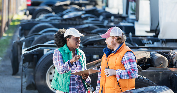 Multi-ethnic workers at a trucking company, standing by a fleet of semi-trucks, conversing. An African-American woman is holding a clipboard, giving directions to her coworker. She is in her 30s and the man is in his 40s.