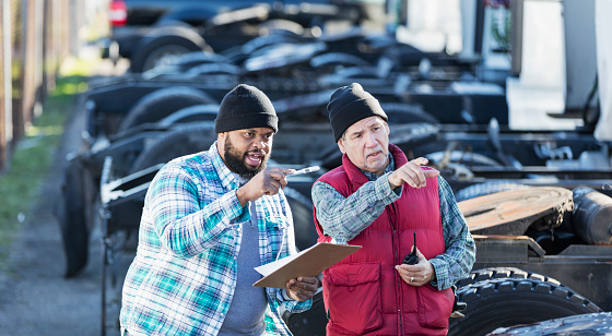 Multi-ethnic workers at a trucking company, standing in front of a fleet of semi-trucks, conversing. An African-American man in his 30s is holding a clipboard, giving directions to his coworker, a senior Hispanic man in his 60s.
