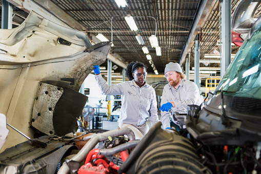 Two multi-ethnic mechanics repairing a semi-truck. They are looking under the hood of the truck, examining the engine. The African-American woman may be a trainee. She is looking and listening to the man with a beard.