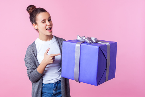Look at this present. Portrait of beautiful teenage brunette girl with bun hairstyle in casual clothes pointing at gift box and winking joyfully at camera. studio shot isolated on pink background