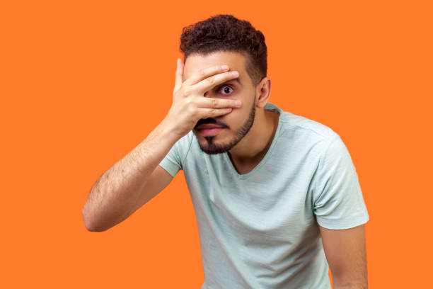portrait of curious brunette man covering face, looking through fingers with big eyes. isolated on orange background - spy secrecy top secret mystery imagens e fotografias de stock