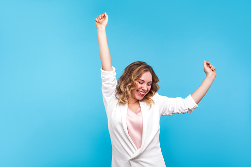 I'm winner! Portrait of ecstatic overjoyed lady with wavy hair in white jacket dancing with raised arms, smiling excitedly, celebrating victory, success. indoor studio shot isolated on blue background