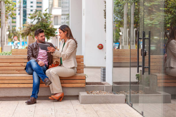 A couple of Latin American colleagues are seen seated outside their office building enjoying the morning sunshine. The are discussing some information using a digital tablet. One of them is more formally attired in a pant suit; while the other is casually dressed. The lady's hair is untied and has been styled with highlights. Horizontal format; Copy space. un par de colegas latinoamericanos son vistos sentados fuera de su edificio de oficinas por la mañana, compartiendo información sobre una tableta digital; la dama está vestida más formalmente, mientras que el hombre está vestido casual. formato horizo