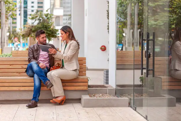 A couple of Latin American colleagues are seen seated outside their office building enjoying the morning sunshine. The are discussing some information using a digital tablet. One of them is more formally attired in a pant suit; while the other is casually dressed. The lady's hair is untied and has been styled with highlights. Horizontal format; Copy space.