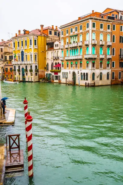 Striped red and white mooring poles on the Grand Canal channel water way. People/ tourists standing with umbrella on rainy day in Venice city, Italy by colorful gothic venetian architecture buildings.
