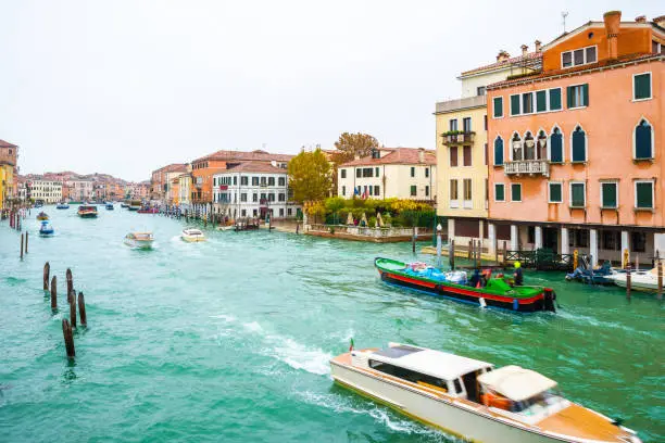 Boats, yachts, vaporettos, water taxis sailing down the Grand Canal waterway between wooden and striped mooring poles and colorful Venetian architecture buildings. November in Venice city, Italy.
