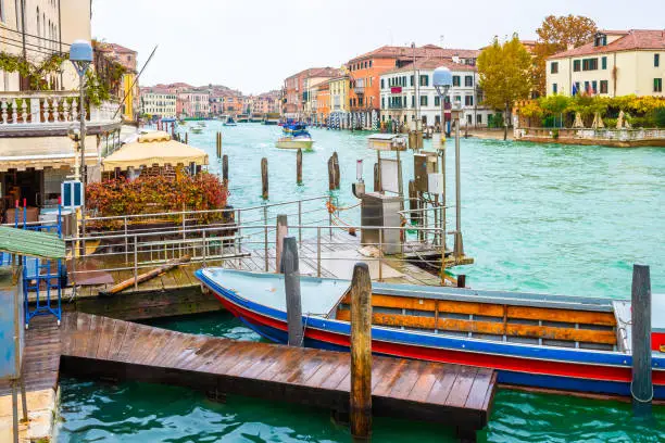 Old boat docked on waterway at wooden mooring poles, other boats/ yachts/ water taxis sailing down the Grand Canal channel, colorful Venetian architecture buildings. November in Venice city, Italy
