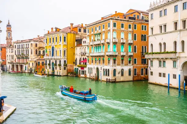 Boats and water taxis sailing and docked on Grand Canal along striped and wooden mooring poles and colorful Venetian architecture buildings. People/ tourists in Venice city Italy on rainy day.