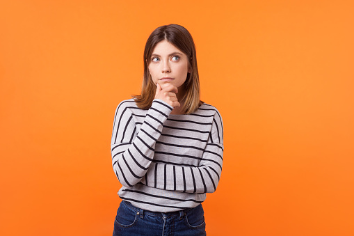Think up plan. Portrait of pensive woman with brown hair in long sleeve striped shirt. indoor studio shot isolated on orange background