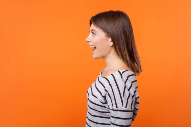 seitenansicht porträt der schönen erstaunte frau mit braunen haaren in langärmeligen gestreiften hemd. indoor-studio-aufnahme isoliert auf orangefarbenem hintergrund - geöffneter mund stock-fotos und bilder