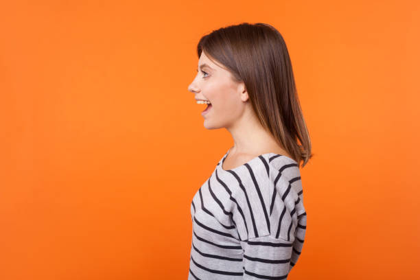 side view portrait of beautiful amazed woman with brown hair in long sleeve striped shirt. indoor studio shot isolated on orange background - mouth open imagens e fotografias de stock