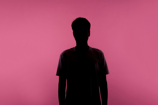 No name, anonymous person hiding face in shadow, human identity. Silhouette portrait of young man in casual T-shirt standing calm with hands down, indoor studio shot, isolated on pink background