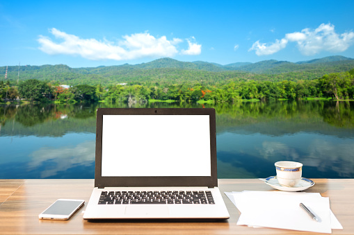 Mockup image of laptop with blank white screen,smart phone and document on wooden table of landscape forested Mountain blue sky background.