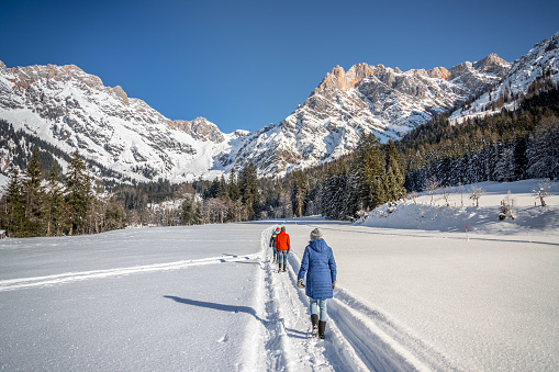 Group of people are walking on a snowy footpath, idyllic winter landscape with stunning mountain range, snowy trees and blue sky