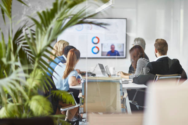 Male and Female Executive Team Video Conferencing with CEO Rear view photographed through window of diverse male and female executive team video conferencing with CEO in office board room. business relationship photos stock pictures, royalty-free photos & images