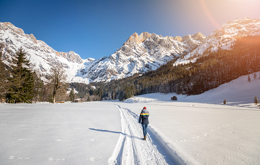 Girl is walking on a snowy footpath, idyllic winter landscape with stunning mountain range, snowy trees and blue sky