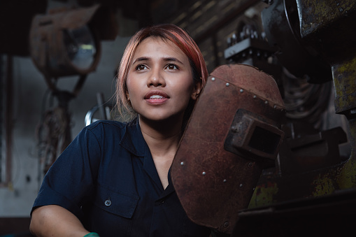Joven mujer asiática aprendiz soldador mirando lejos de la cámara y tomar un descanso del trabajo dentro de la fábrica de talleres de metal photo