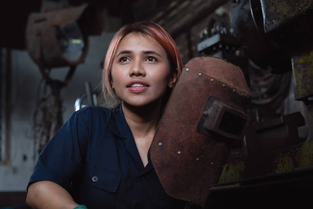 Young Asian female apprentice welder looking away from camera and taking a break from work inside metal workshop factory stock photo