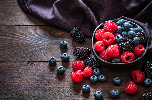 High angle view of a black bowl filled with fresh organic berries like raspberries, blackberries and blueberries on a rustic wooden table.  Low key DSLR photo taken with Canon EOS 6D Mark II and Canon EF 24-105 mm f/4L