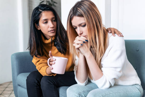 una mujer muy joven apoyando y consolando a su triste amiga mientras se sienta en el sofá en casa. - reconfortante fotografías e imágenes de stock