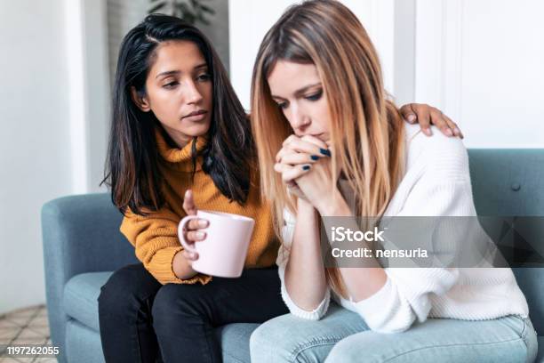 Pretty Young Woman Supporting And Comforting Her Sad Friend While Sitting On The Sofa At Home Stock Photo - Download Image Now