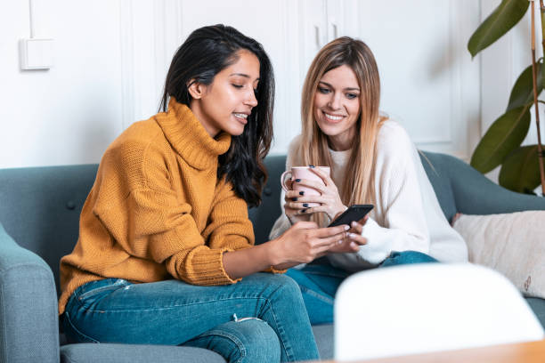 istock Two pretty young women using mobile phone while drinking coffee on sofa at home. 1197262065