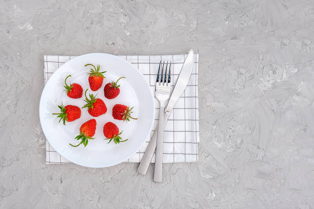 Red ripe strawberries berry on white plate, cutlery and red alarm clock on gray stone background table. Top view, flat lay, copy space Template. Concept diet and detox time or summer menu time