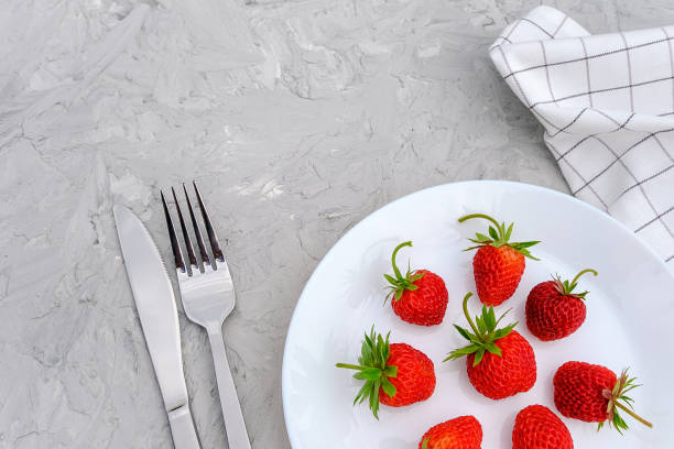 Red ripe strawberries berry on white plate, cutlery and red alarm clock on gray stone background table. Top view, flat lay, copy space Template. Concept diet and detox time or summer menu time