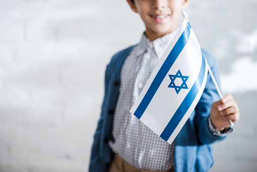 Little girl is holding flag of Israel in hand and is looking at camera in front of white background.