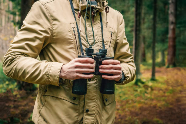 um homem em um chapéu e em um verde e em bege uniformes prende binóculos e olha na distância, guarda florestal que presta atenção ao território, a proteção da reserva - 4679 - fotografias e filmes do acervo