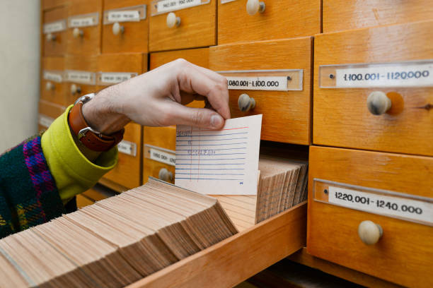поиск информации в базе данных. мужчина держит в руках бумажную карточку с информацией. деревянный шкаф в офисе или библиотеке. секретные д� - furniture office old warehouse стоковые фото и изображения