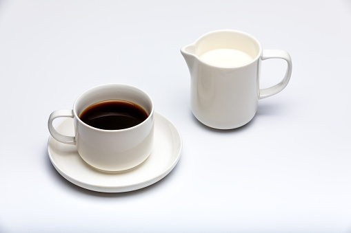 Creamer with milk and cup of coffee on white background.