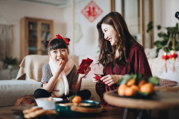 Lovely daughter enjoying traditional snacks while helping her mother to prepare red envelops (lai see) at home for Chinese New Year Lovely daughter enjoying traditional snacks while helping her mother to prepare red envelops (lai see) at home for Chinese New Year luck photos stock pictures, royalty-free photos & images
