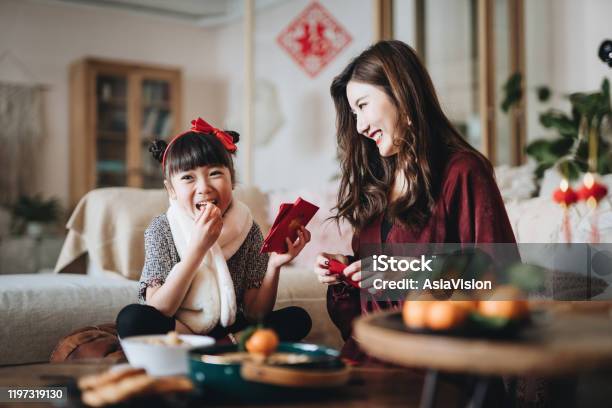 Lovely Daughter Enjoying Traditional Snacks While Helping Her Mother To Prepare Red Envelops At Home For Chinese New Year Stock Photo - Download Image Now