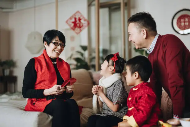 Photo of Cute little granddaughter and grandson dressed in red traditional Chinese costume greeting to grandparents and receives red envelops (lai see) joyfully in Chinese New Year